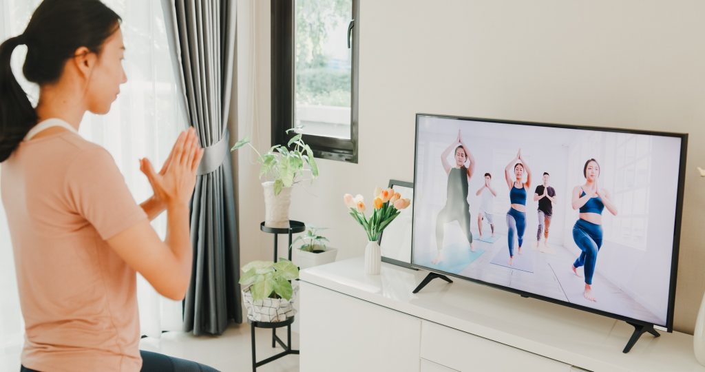 woman in sportswear site on mat in the floor virtual fitness class with group of people on a video.