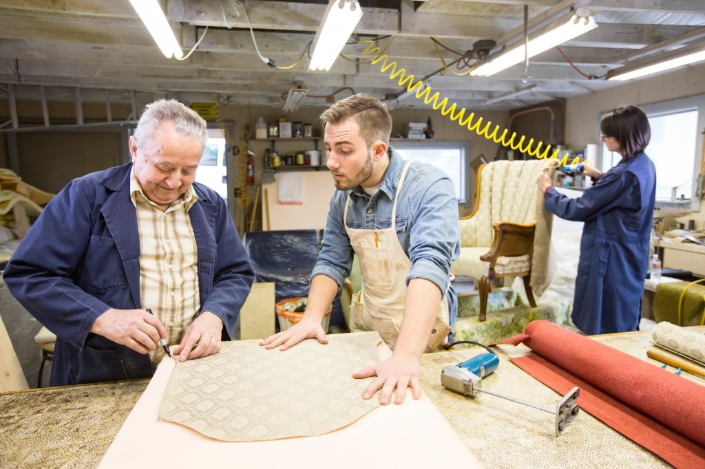Young Caucasian man learning the art of upholstery from a senior male upholsterer.